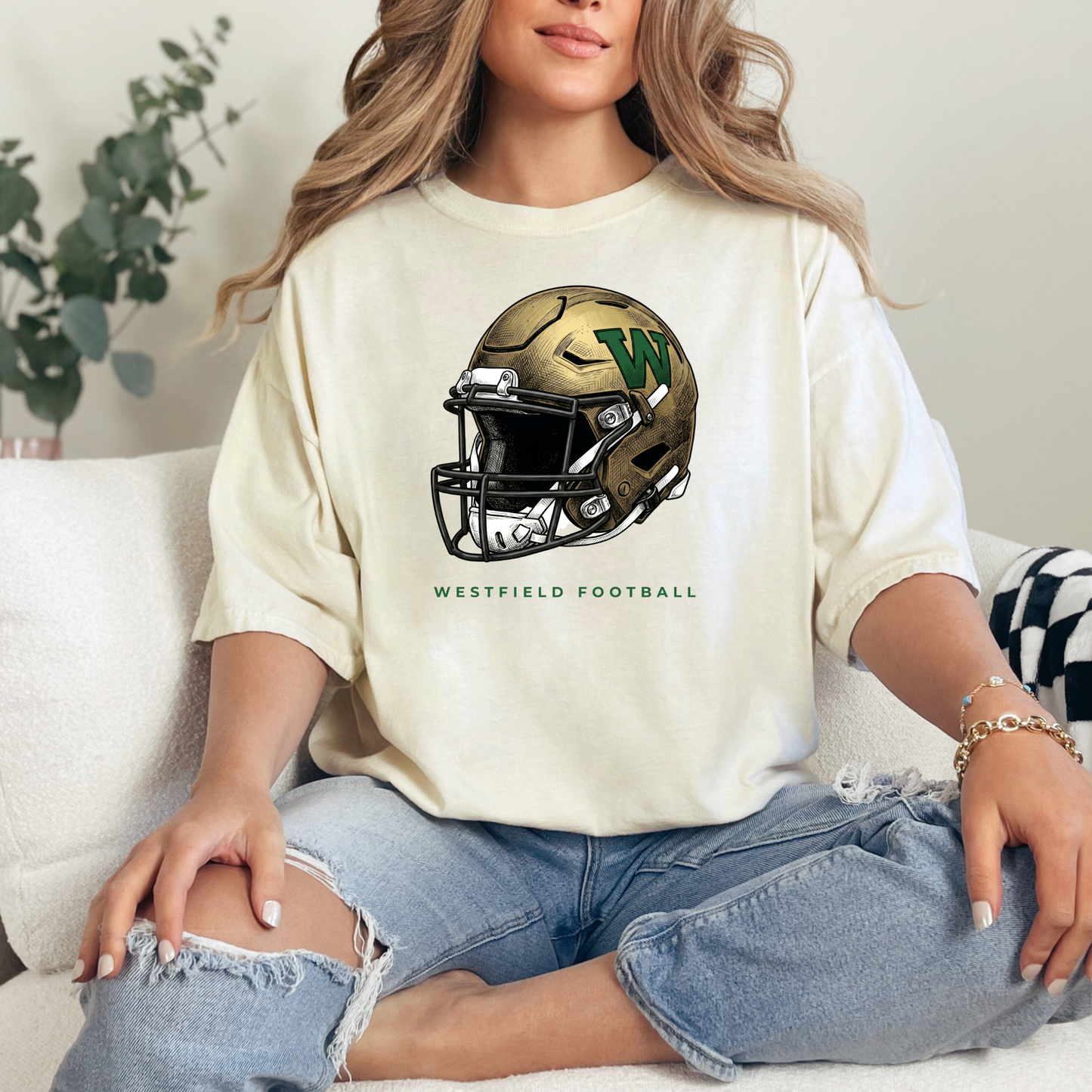 A person wearing a cream-colored t-shirt featuring a gold and green Westfield football helmet design with the text 'WESTFIELD FOOTBALL', sitting on a couch.