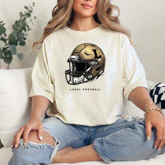 A person sitting on a couch wearing a cream-colored t-shirt featuring a detailed gold football helmet graphic with 'LAPEL FOOTBALL' text.
