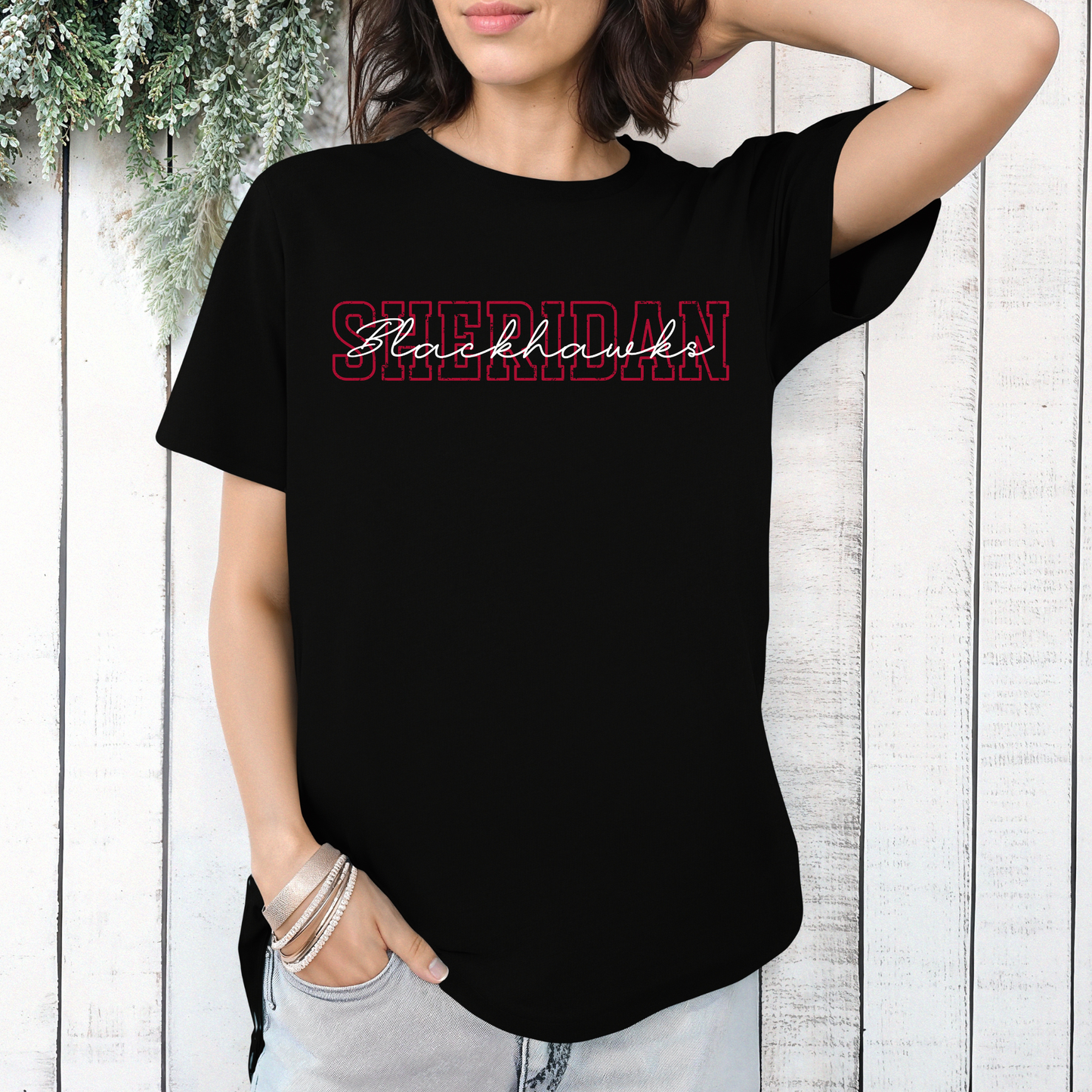 Woman wearing a black graphic t-shirt featuring a layered, vintage-style Sheridan Blackhawks block and script typography design, styled against a white farmhouse-style wooden wall.
