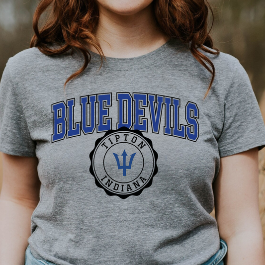 Woman wearing a grey graphic t-shirt featuring a collegiate-style Tipton Blue Devils and Indiana seal graphic with a trident icon.