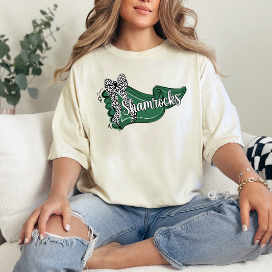 A person sitting on a couch wearing a cream-colored t-shirt featuring a green banner design with 'Shamrocks' text and a leopard-print bow.