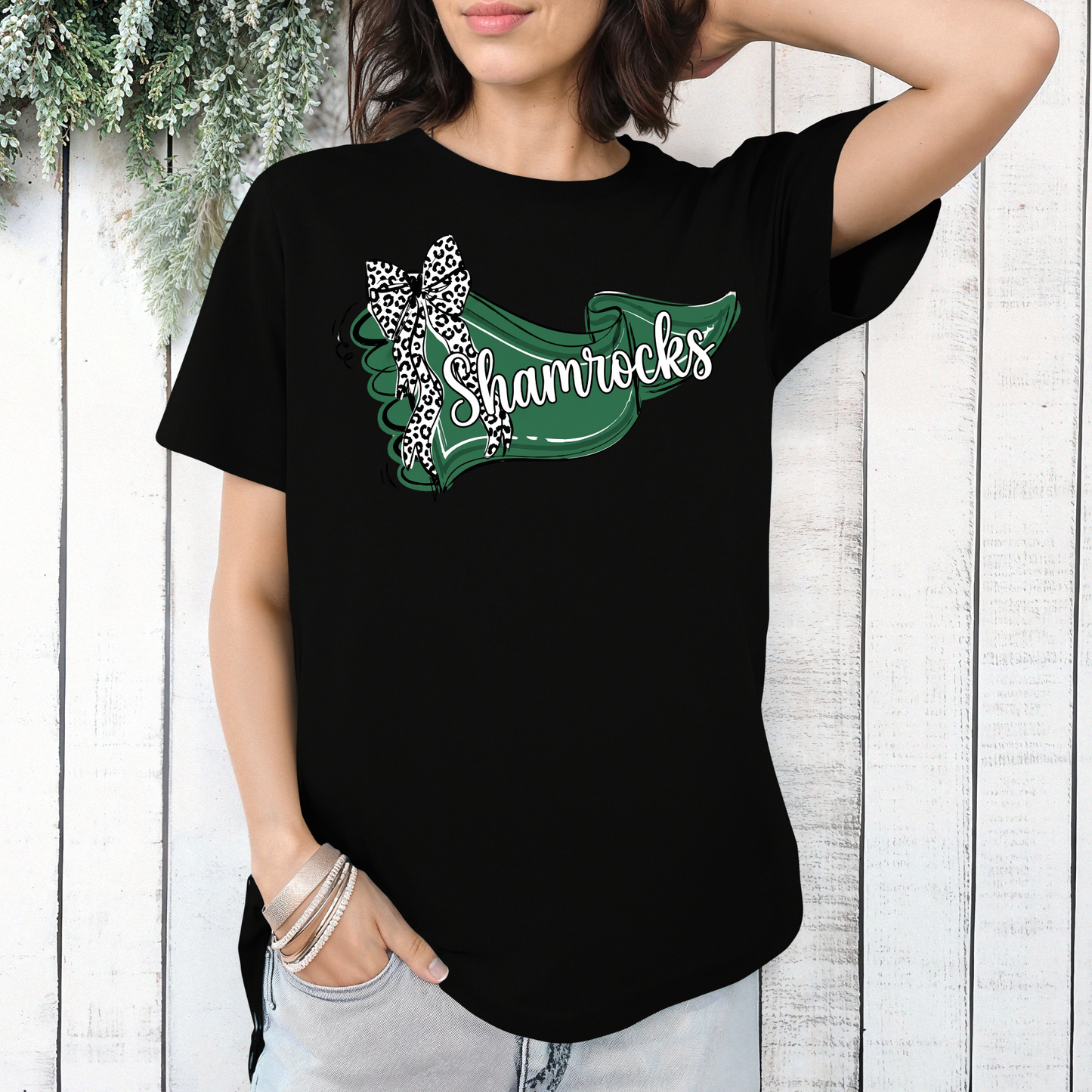 A person wearing a black t-shirt featuring a green banner design with 'Shamrocks' text and a leopard-print bow, posed in front of a white wooden wall.