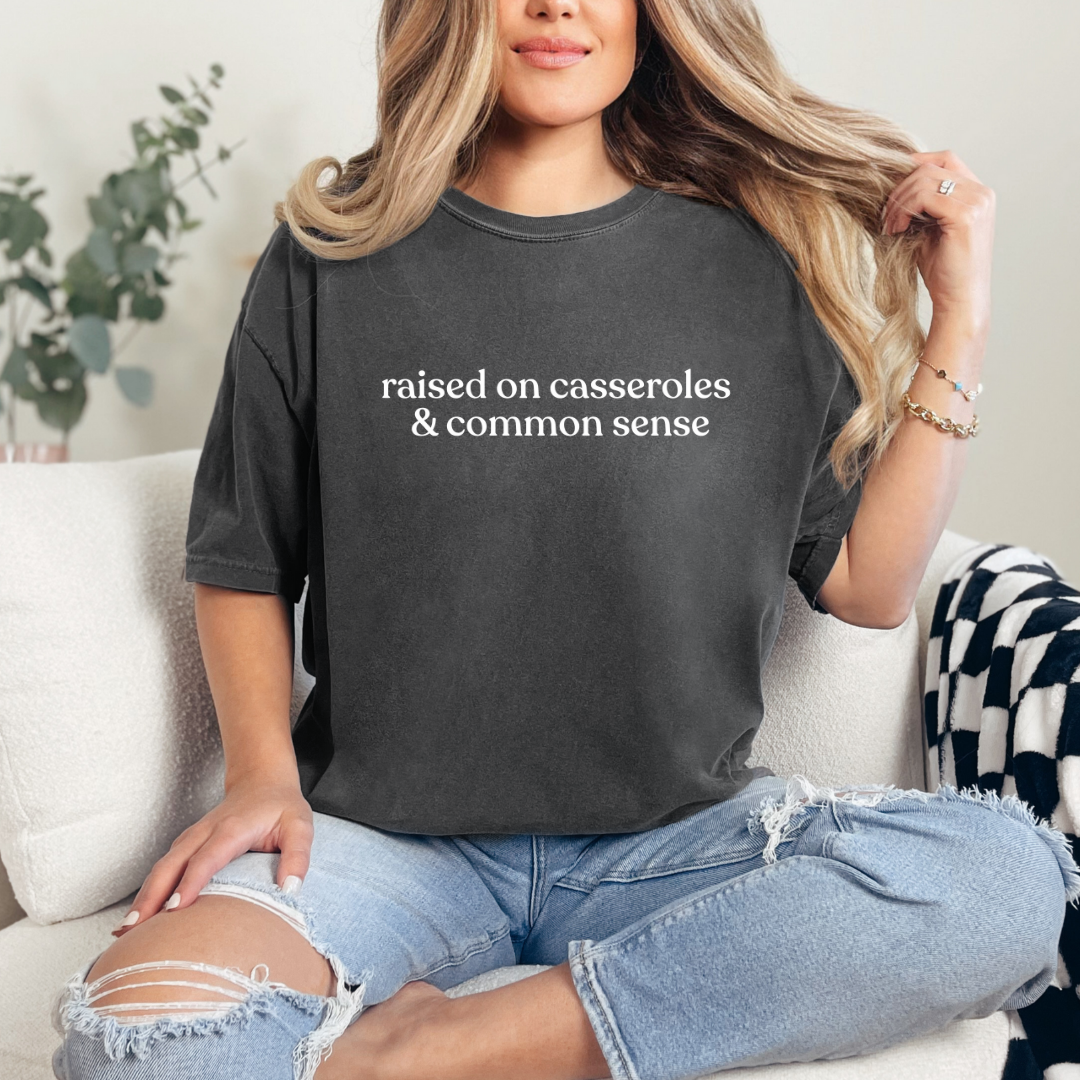 A woman sitting casually on a sofa wearing a pepper Comfort Colors T-shirt with the phrase ‘raised on casseroles & common sense’ printed in black. Cozy, relaxed home setting.