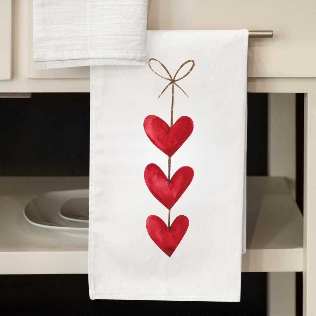 Flour sack kitchen tea towel hanging in front of a kitchen sink on a drawer handle featuring red colorful hearts on a string.