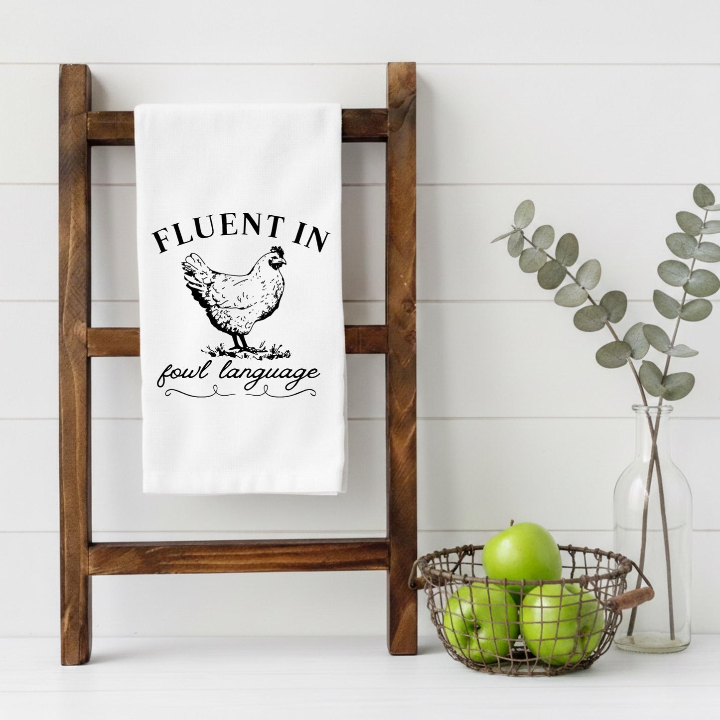 White towel with chicken graphic and text on a wooden ladder against a white wall.