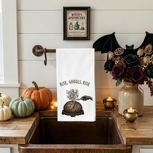 A white kitchen towel with a black gothic illustration and the text 'RISE, Ghouls, RISE' hangs over a rustic copper sink. The scene is styled for Halloween with pumpkins, a black bat decoration, a vase of dark roses, and candles on a white shiplap wall.