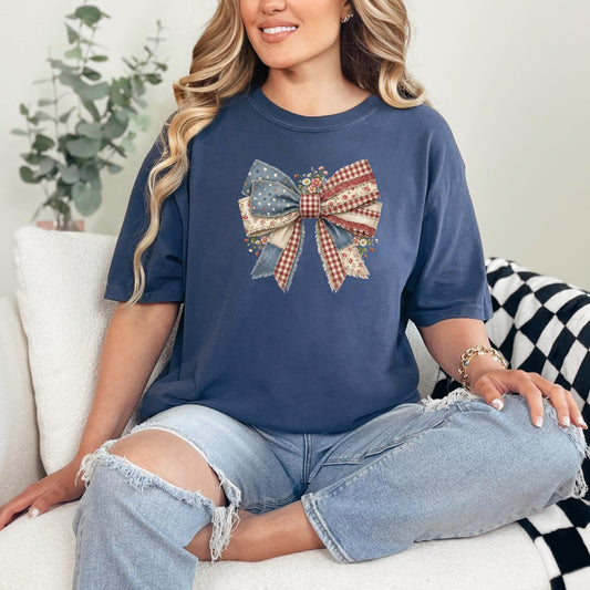 A woman sitting on a white couch wearing a navy blue t-shirt with a large patriotic bow graphic on the front, paired with distressed light-wash jeans.