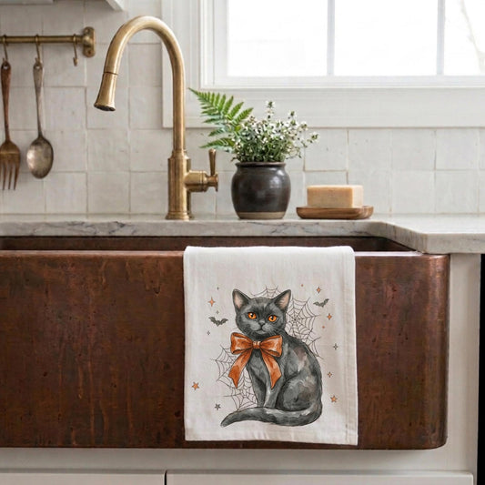 A white kitchen towel with a printed illustration of a black cat wearing an orange bow and surrounded by spiderwebs, hanging over the front of a copper farmhouse sink in a kitchen setting.