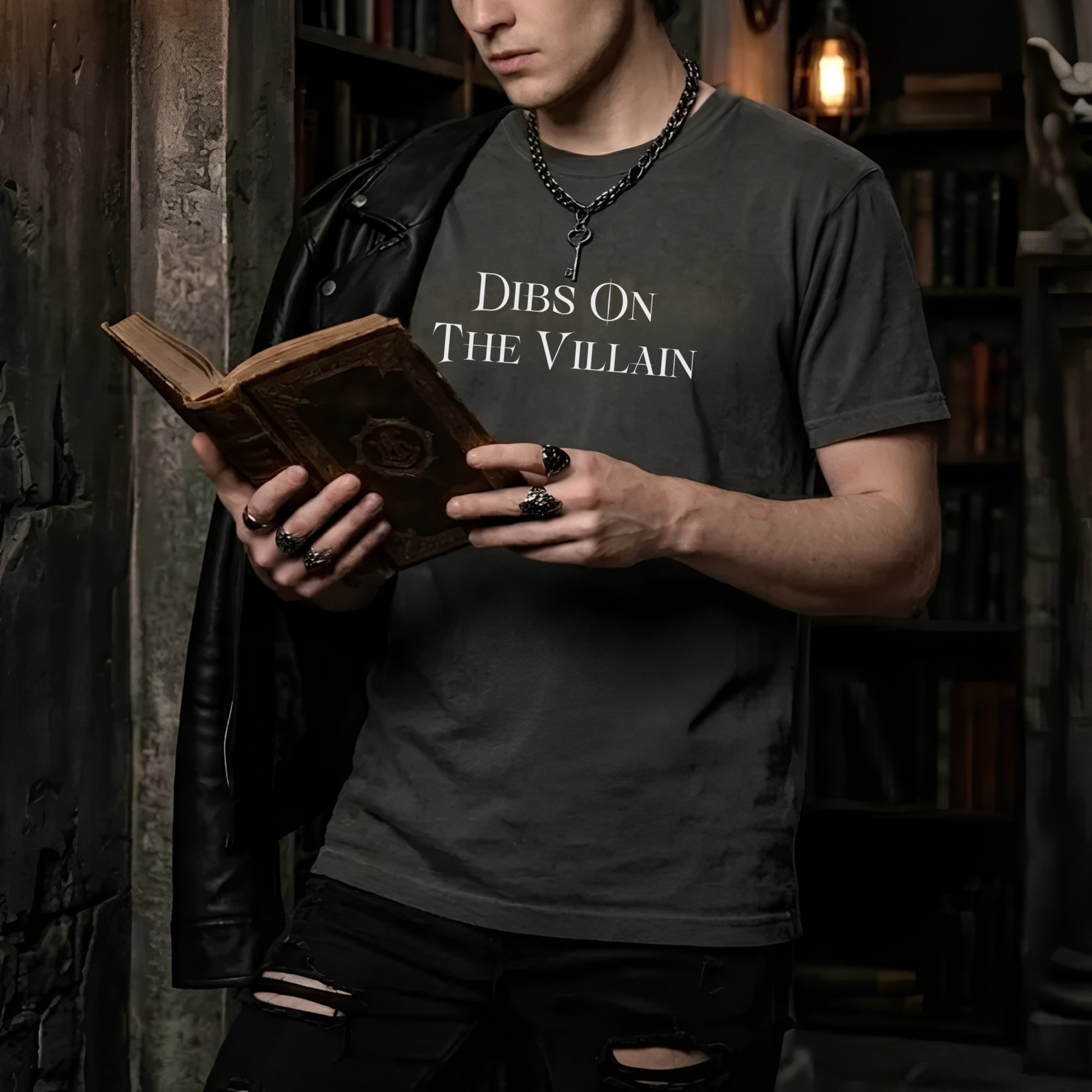 Man wearing Dibs On The Villain shirt while reading a fantasy novel in a dark academia library setting