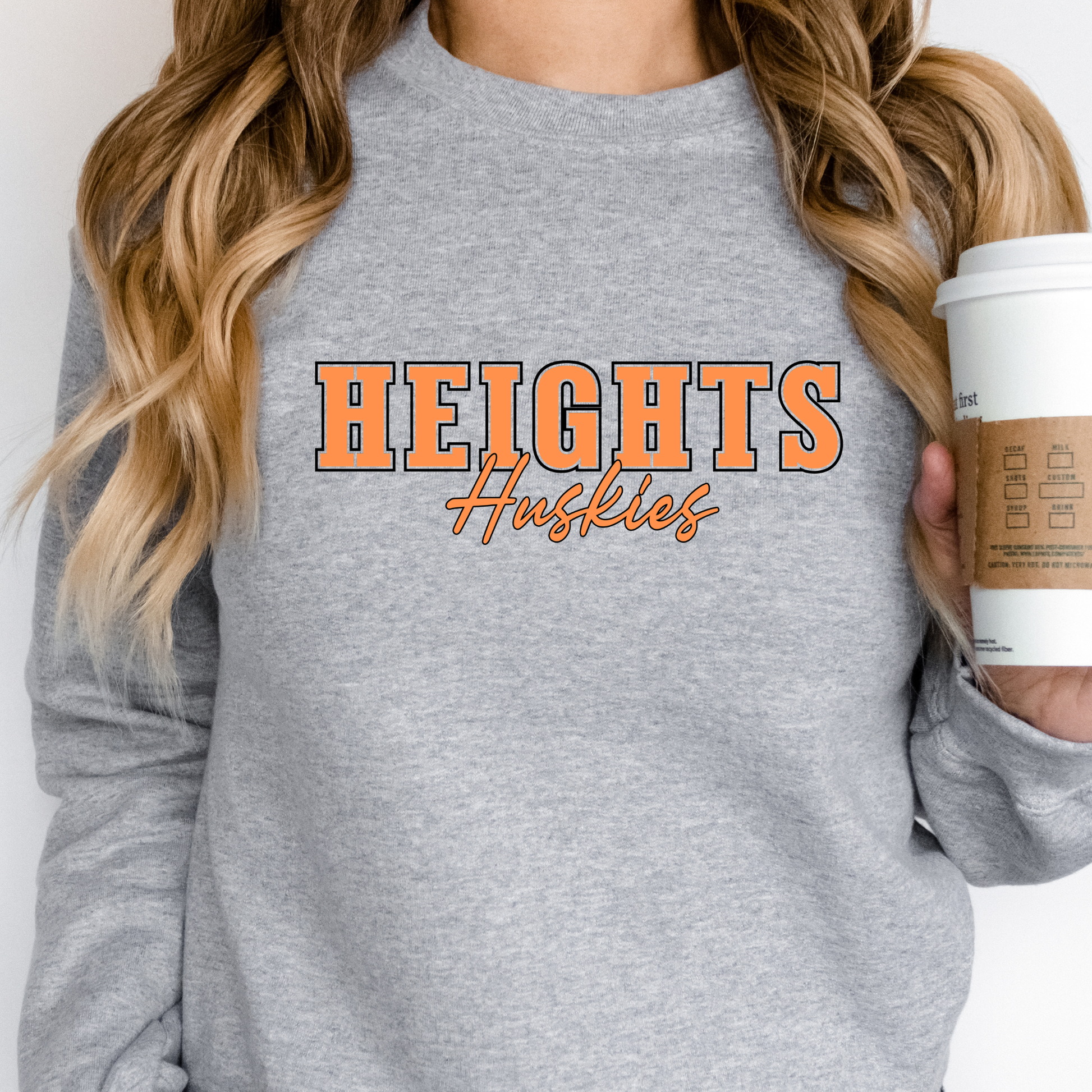 Woman wearing a heather grey Heights Huskies Script Spirit Crewneck Sweatshirt featuring bold orange varsity lettering with Huskies script design in school colors on a comfortable crewneck sweatshirt.