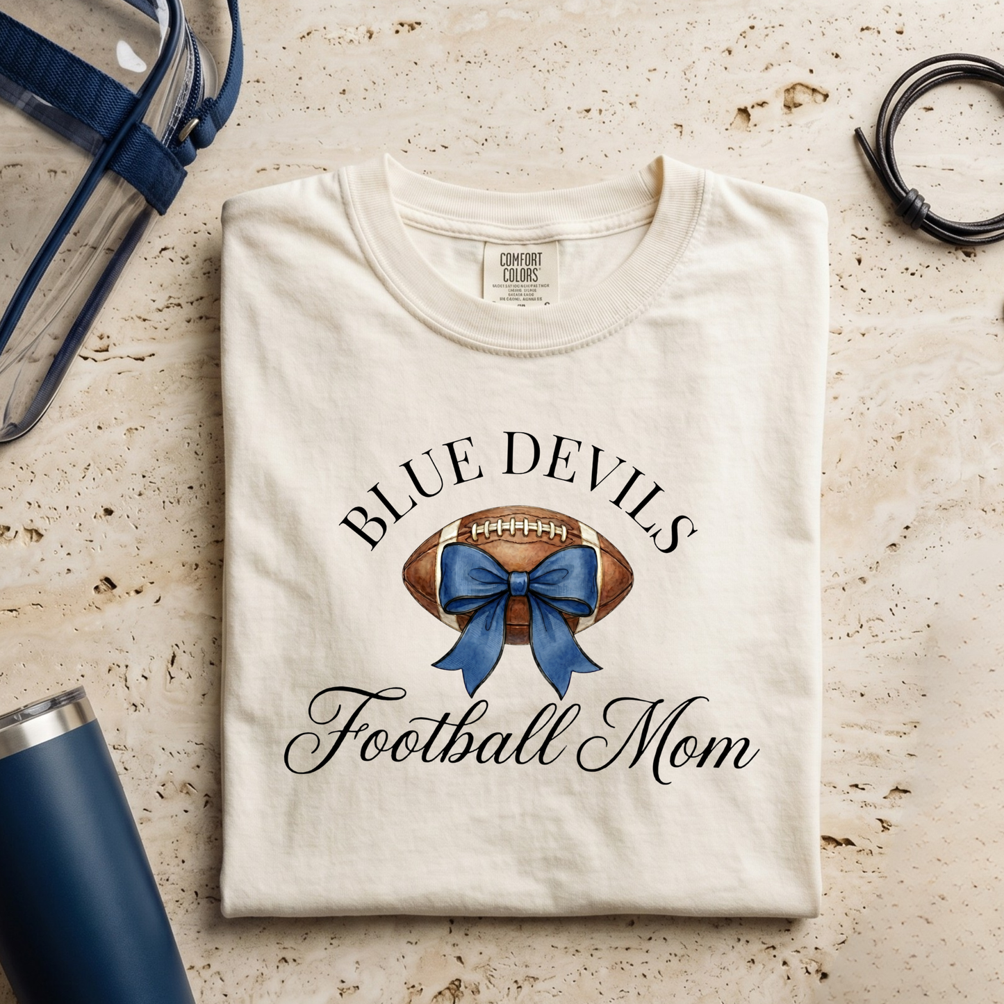 Folded t-shirt with 'Blue Devils Football Mom' design on a textured surface with accessories.