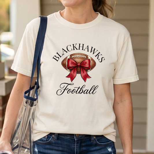 Close-up of Sheridan Blackhawks Football shirt with watercolor football and red bow design on a cream Comfort Colors graphic tee