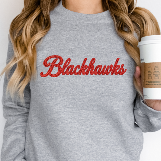 Woman wearing a gray sweatshirt with 'Blackhawks' in red text, holding a coffee cup. Sheridan Blackhawks faux embroidered crewneck sweatshirt in black featuring red script school spirit design.