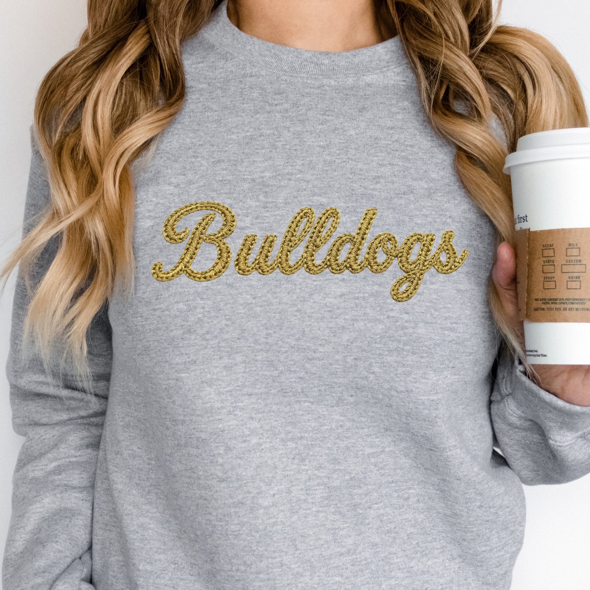 Woman wearing a grey Lapel Bulldogs Faux Embroidered Crewneck Sweatshirt featuring a gold faux embroidered Bulldogs script design for school spirit apparel.