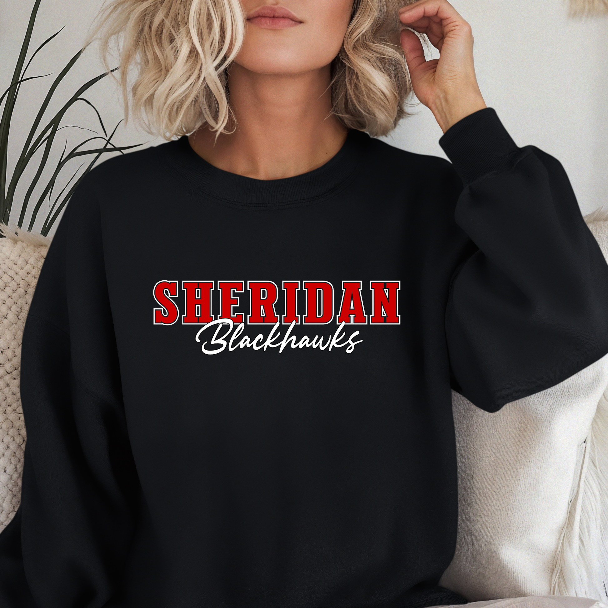 Woman wearing a Sheridan Blackhawks Script Spirit Shirt featuring bold red varsity Sheridan lettering with Blackhawks script design on a unisex crewneck sweatshirt.