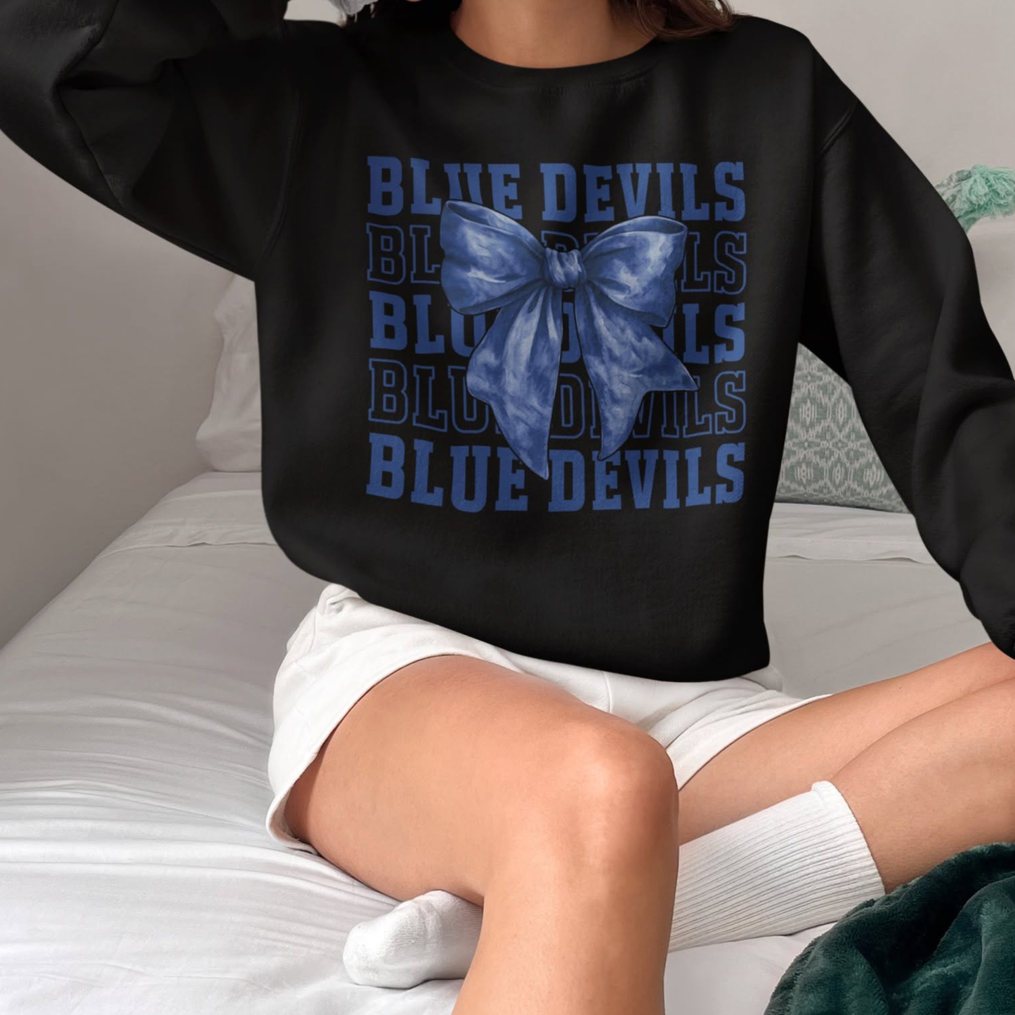 Person wearing a black sweatshirt with 'Blue Devils' text and a blue bow, sitting on a bed.