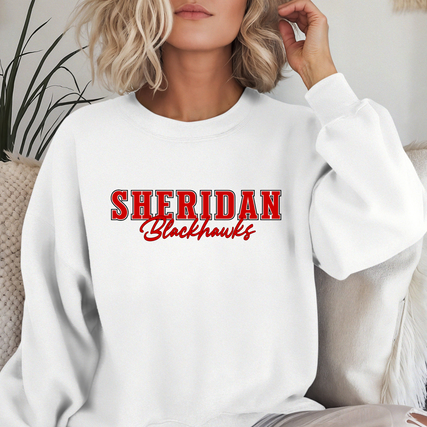 Woman wearing a Sheridan Woman wearing a Blackhawks Script Spirit Shirt featuring bold red varsity Sheridan lettering with Blackhawks script design on a white unisex crewneck sweatshirt.