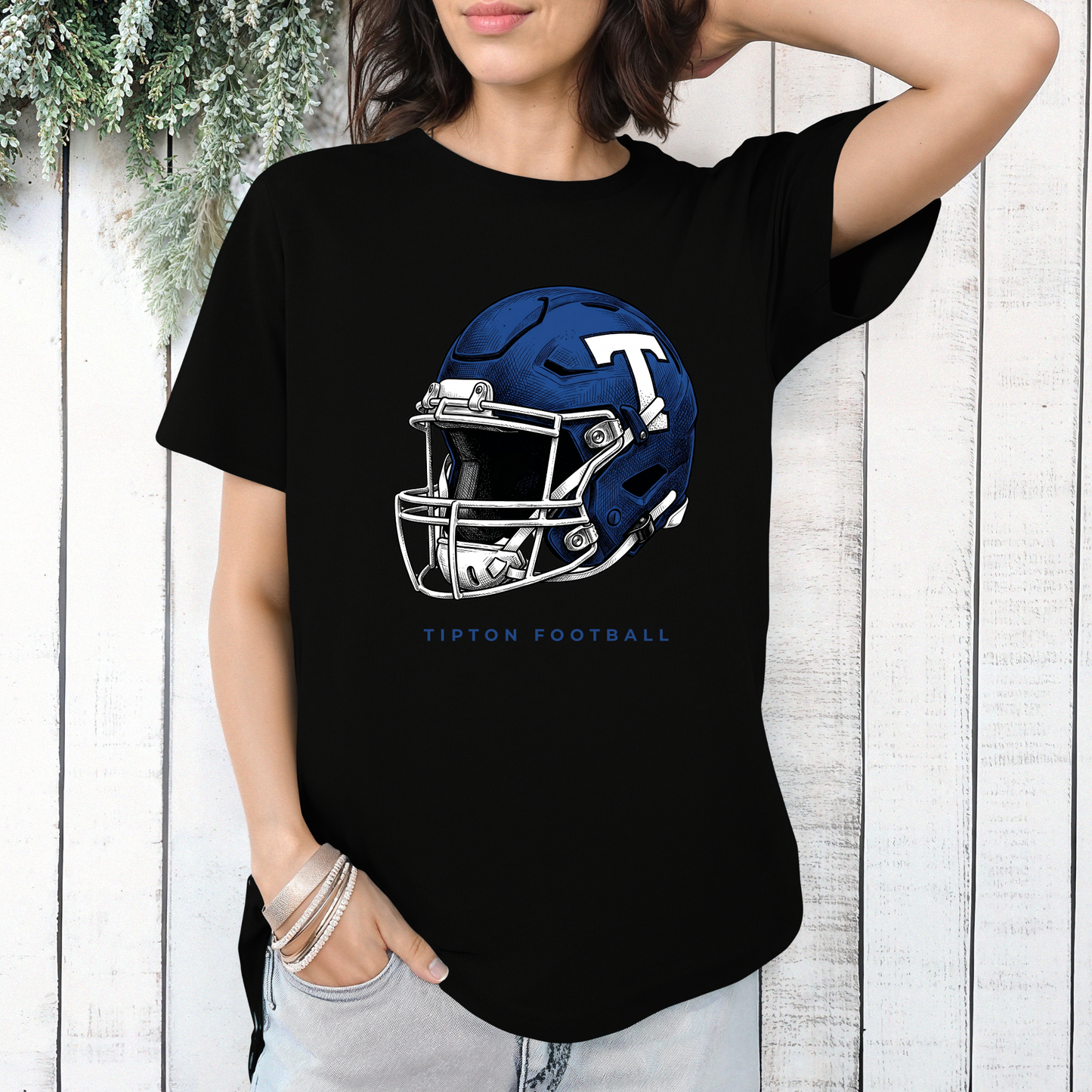 Woman wearing a black graphic t-shirt featuring a detailed Tipton Blue Devils football helmet illustration, styled against a white wooden wall.