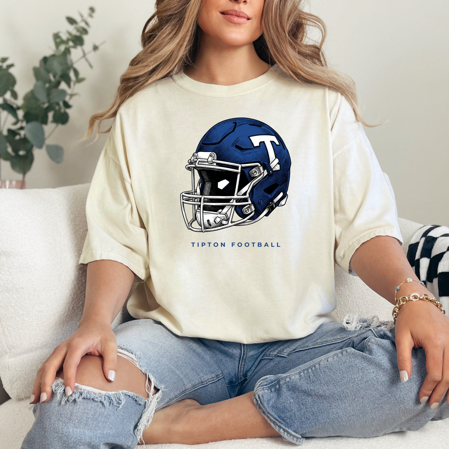 Woman sitting on a couch wearing a cream-colored graphic t-shirt featuring a detailed Tipton Blue Devils football helmet illustration.