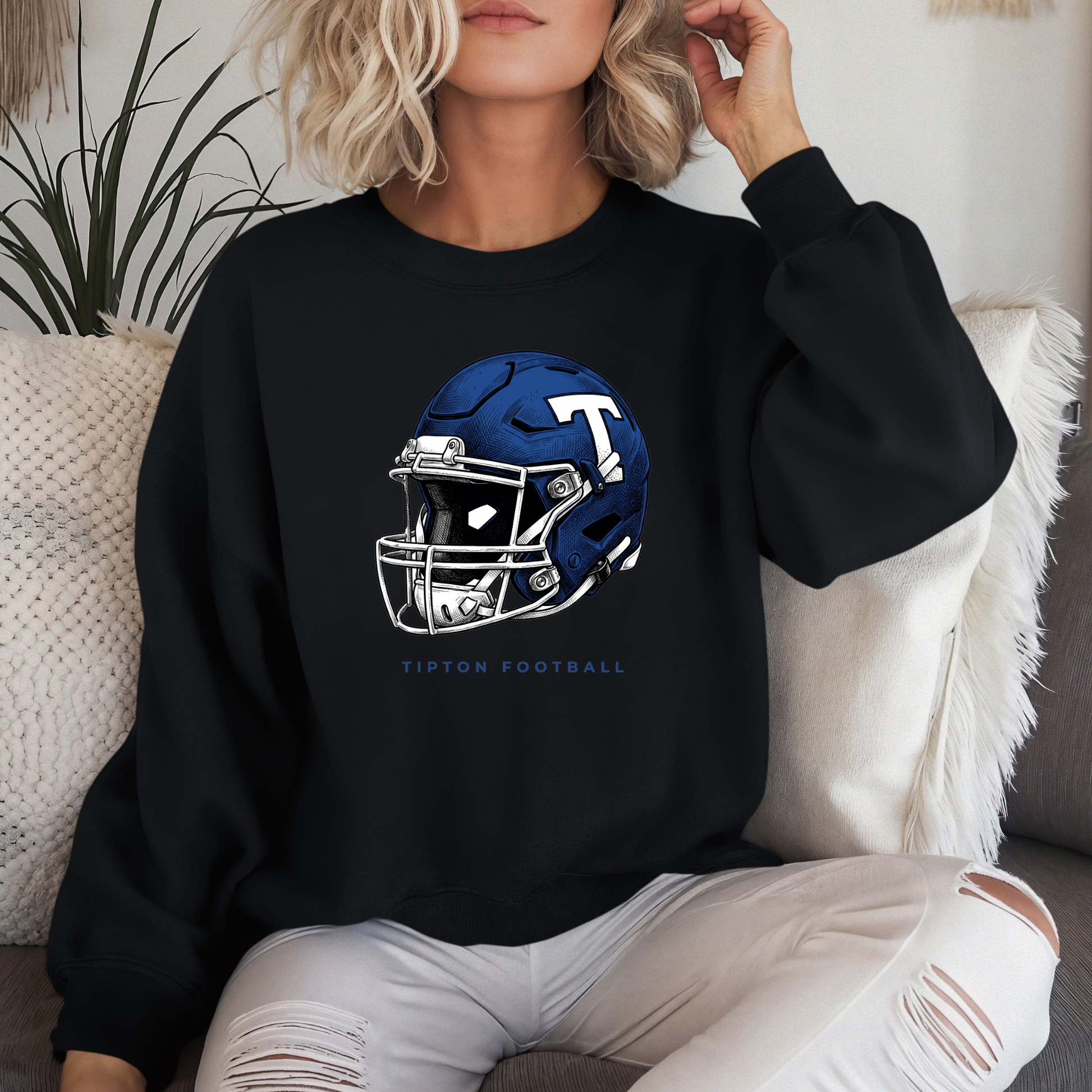 Woman sitting on a couch wearing a black crewneck sweatshirt featuring a detailed Tipton Blue Devils football helmet illustration.
