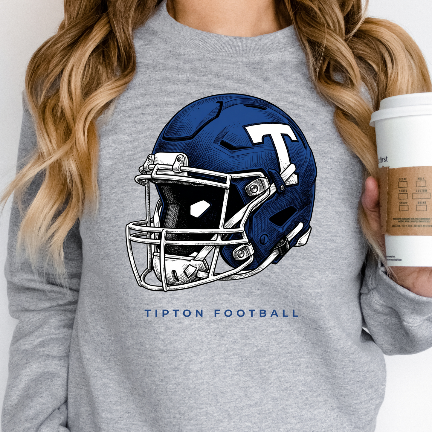 Woman holding a coffee cup, wearing a grey crewneck sweatshirt featuring a detailed Tipton Blue Devils football helmet illustration.