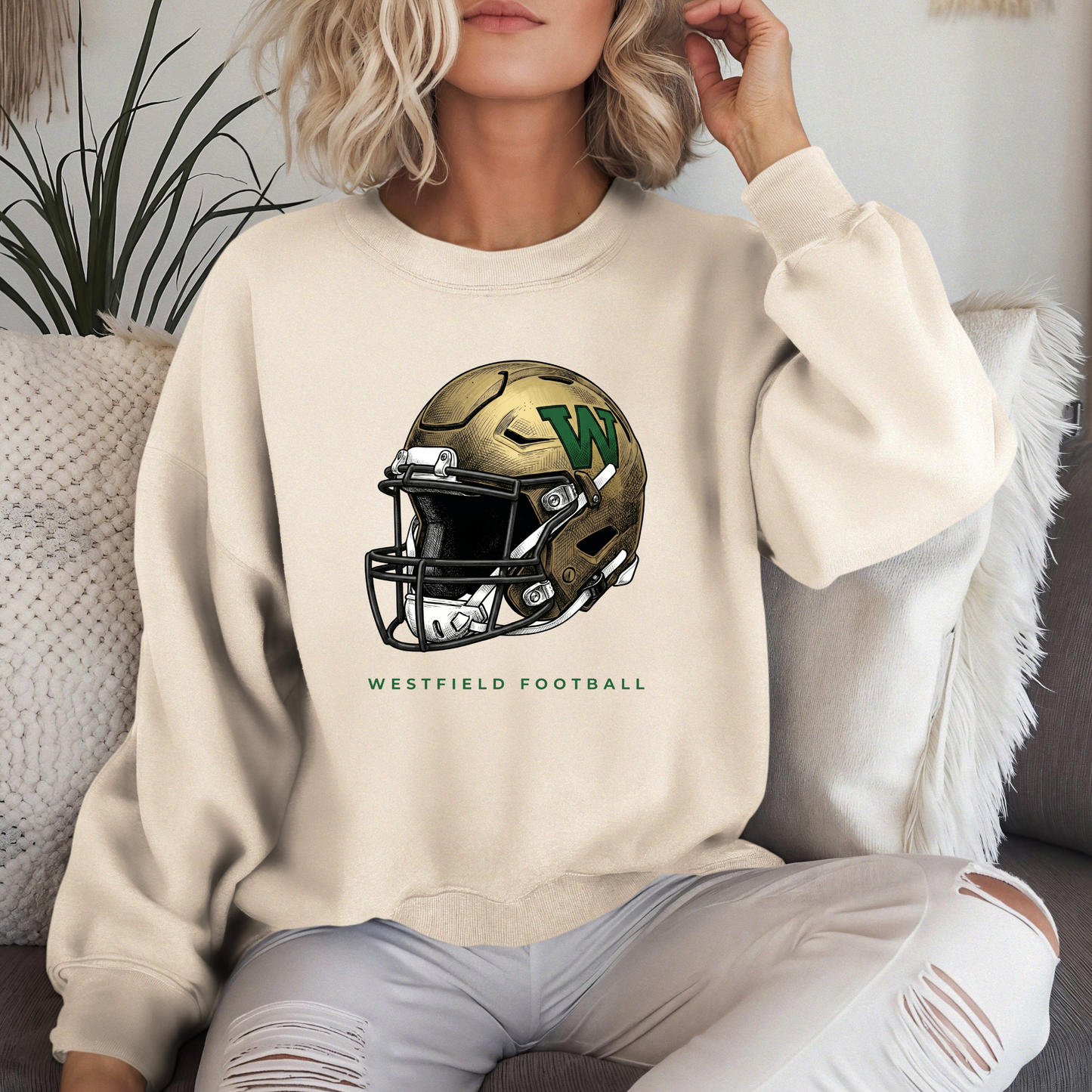 A person lounging in a cream-colored crewneck sweatshirt featuring a gold and green Westfield football helmet design with the text 'WESTFIELD FOOTBALL'.