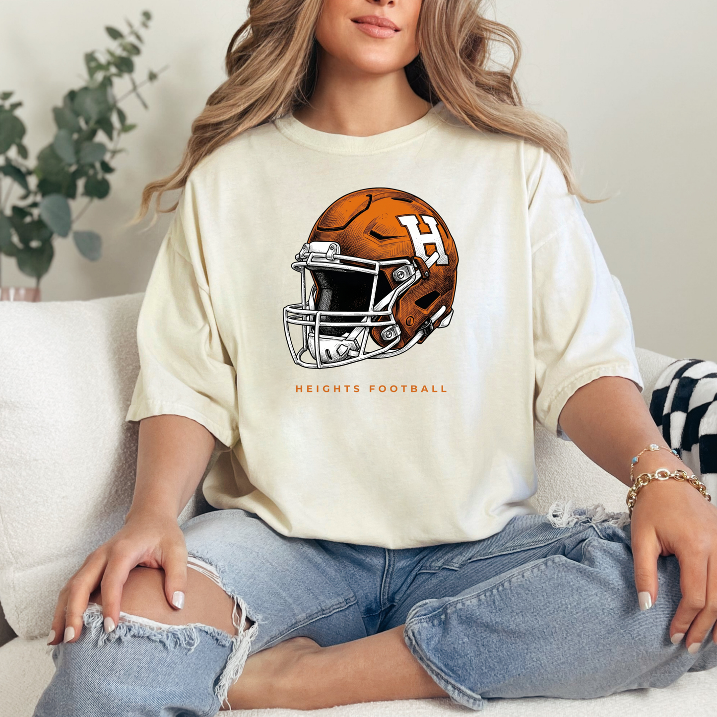 Woman sitting on a couch wearing a cream-colored graphic t-shirt featuring an orange and white Hamilton Heights football helmet design with the text 'HEIGHTS FOOTBALL'.