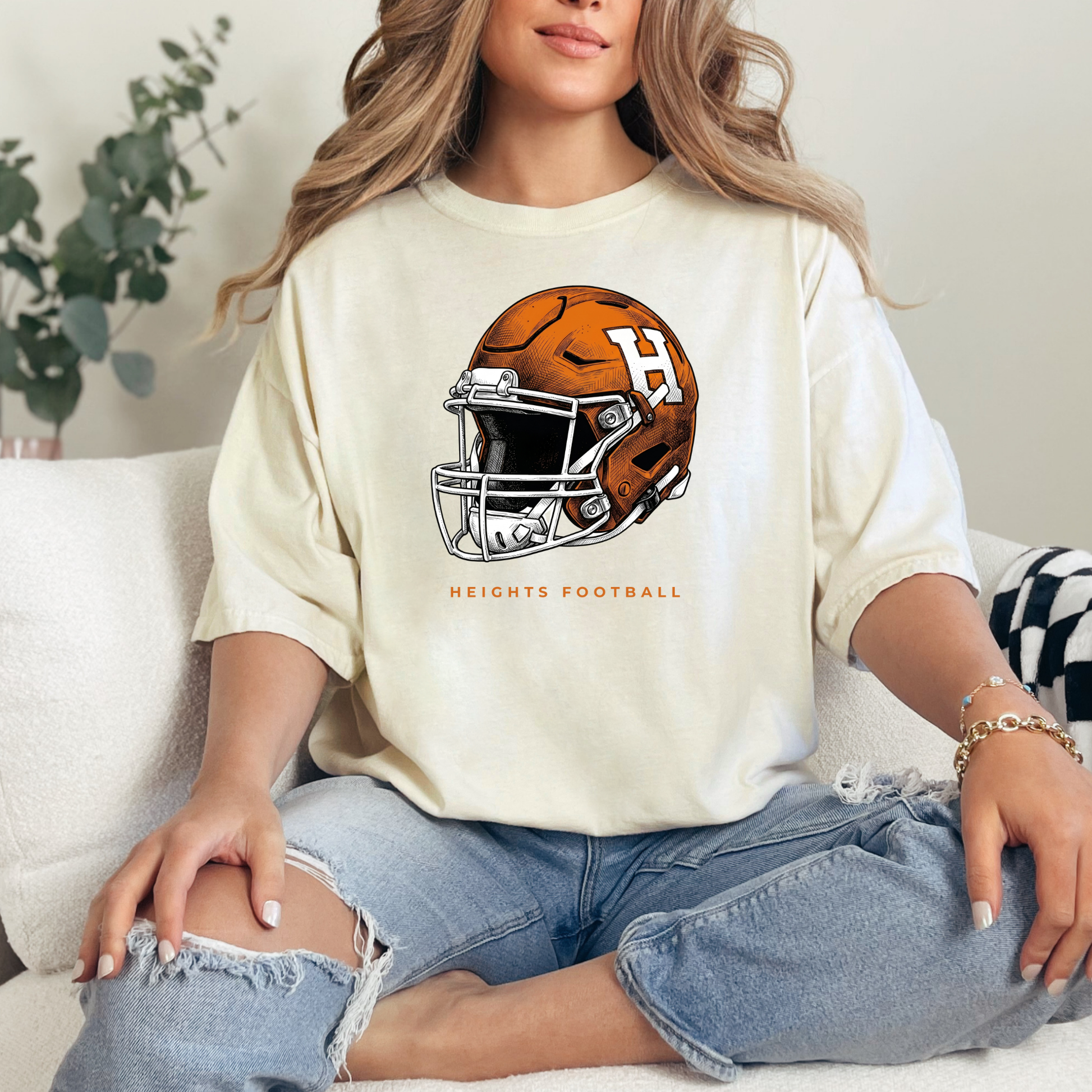 Woman sitting on a couch wearing a cream-colored graphic t-shirt featuring an orange and white Hamilton Heights football helmet design with the text 'HEIGHTS FOOTBALL'.