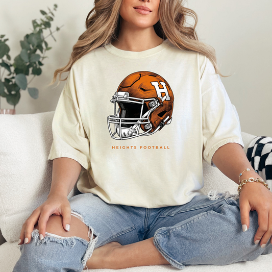 Woman sitting on a couch wearing a cream-colored graphic t-shirt featuring an orange and white Hamilton Heights football helmet design with the text 'HEIGHTS FOOTBALL'.