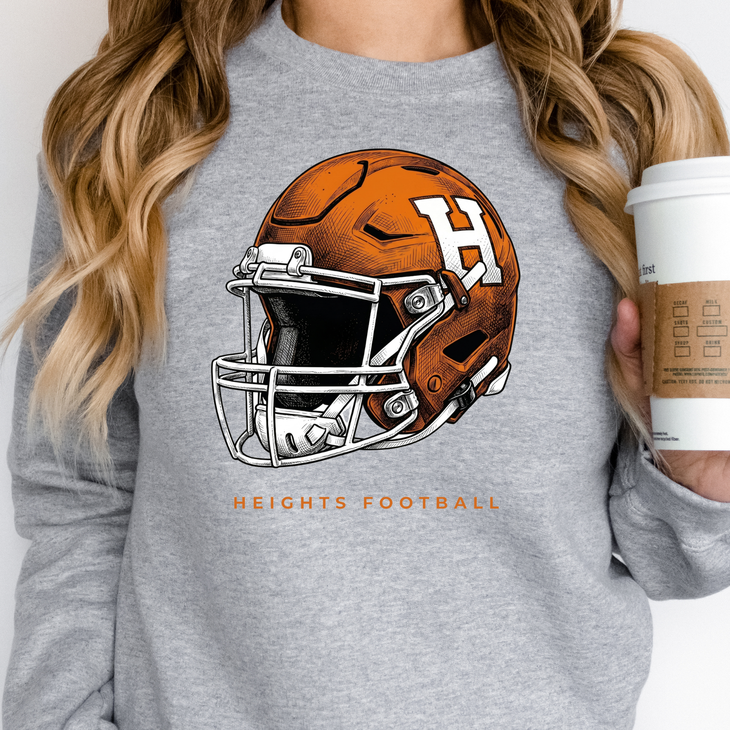 A person wearing a grey crewneck sweatshirt featuring an orange and white Hamilton Heights football helmet design, holding a coffee cup.