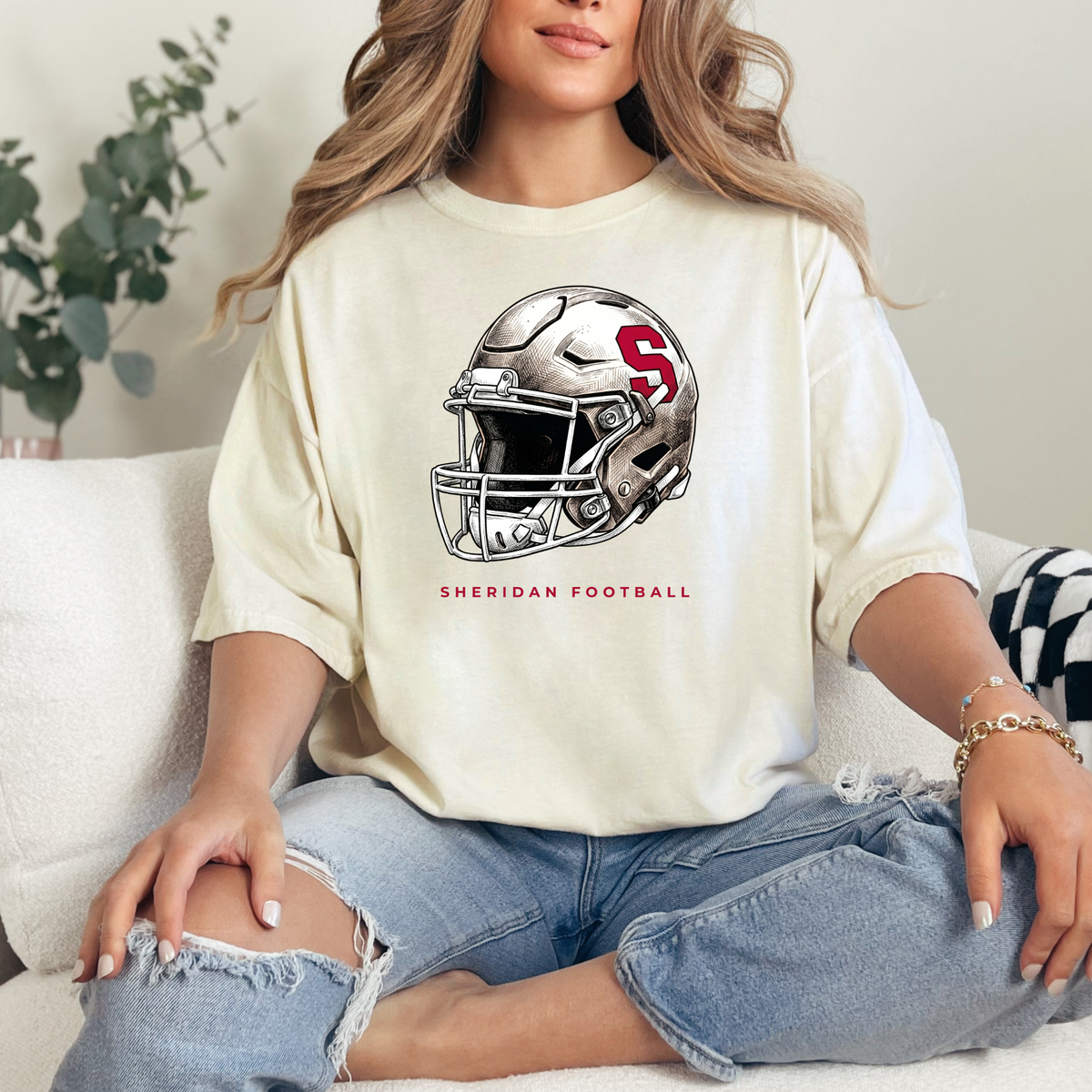 Woman wearing a cream-colored premium graphic t-shirt with a modern Sheridan Blackhawks football helmet design, styled in a relaxed, comfortable fit.