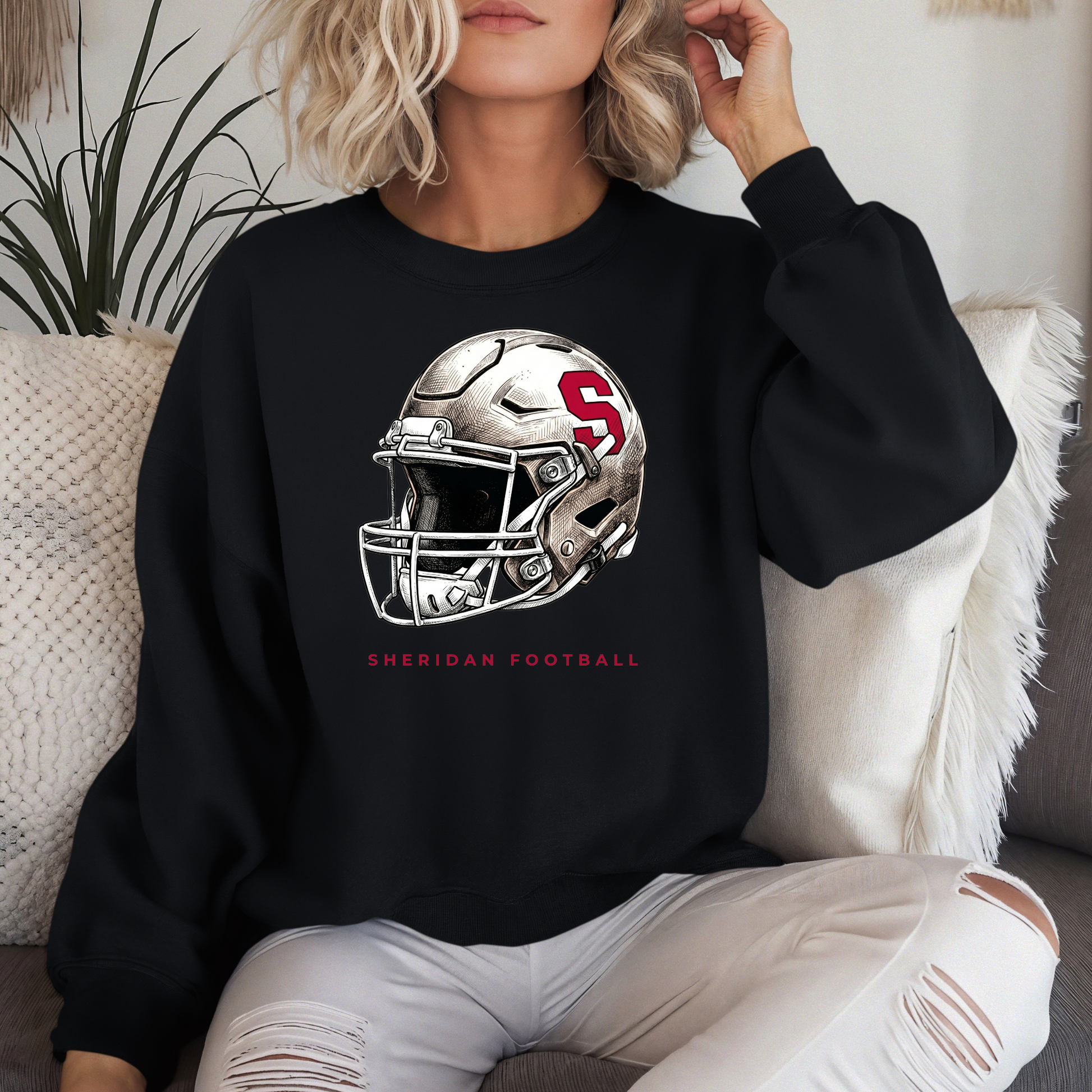 Woman wearing a black crewneck sweatshirt with a modern Sheridan Blackhawks football helmet design, sitting on a couch with white pillows.