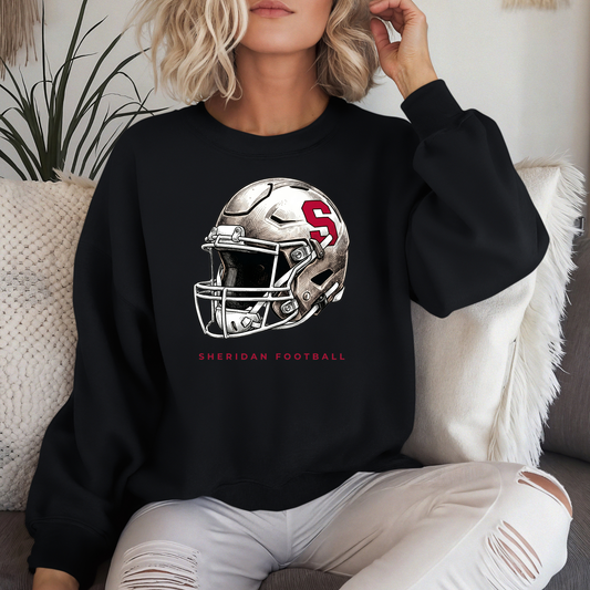 Woman wearing a black crewneck sweatshirt with a modern Sheridan Blackhawks football helmet design, sitting on a couch with white pillows.