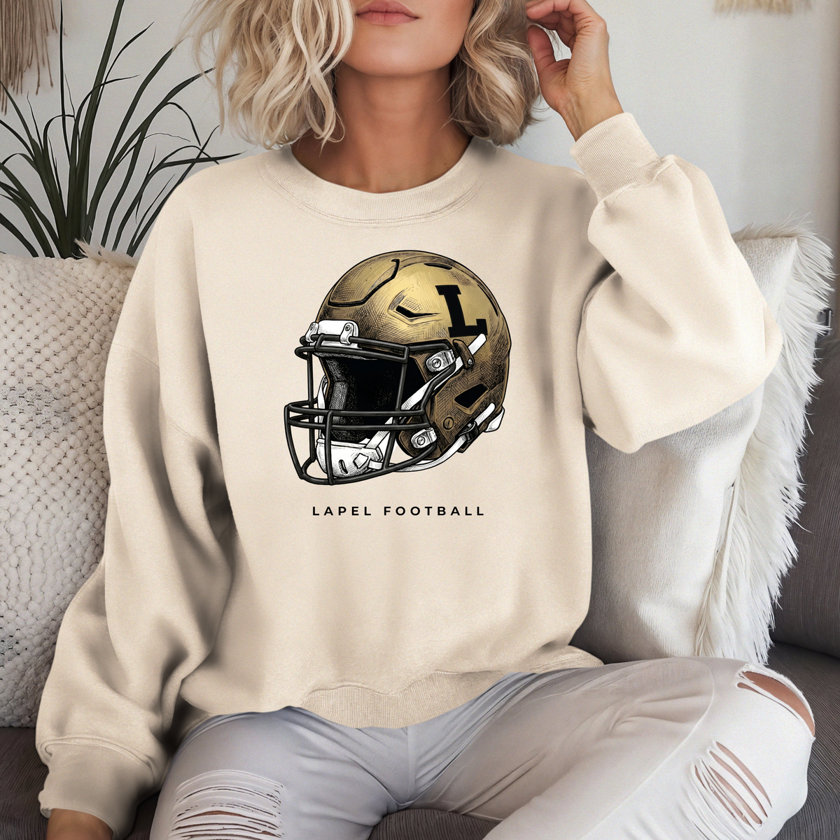 A person sitting on a couch wearing a cream-colored crewneck sweatshirt featuring a detailed gold football helmet graphic with 'LAPEL FOOTBALL' text.