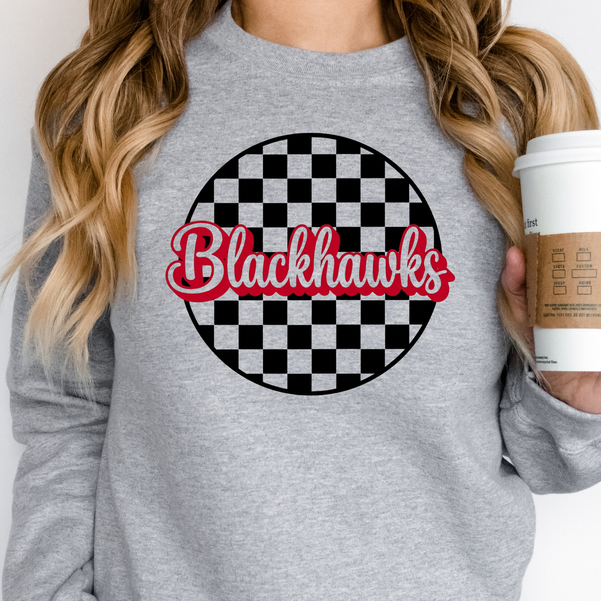 A woman wearing a Sheridan Blackhawks checkered crewneck sweatshirt featuring red script lettering over a black and gray checkerboard design 