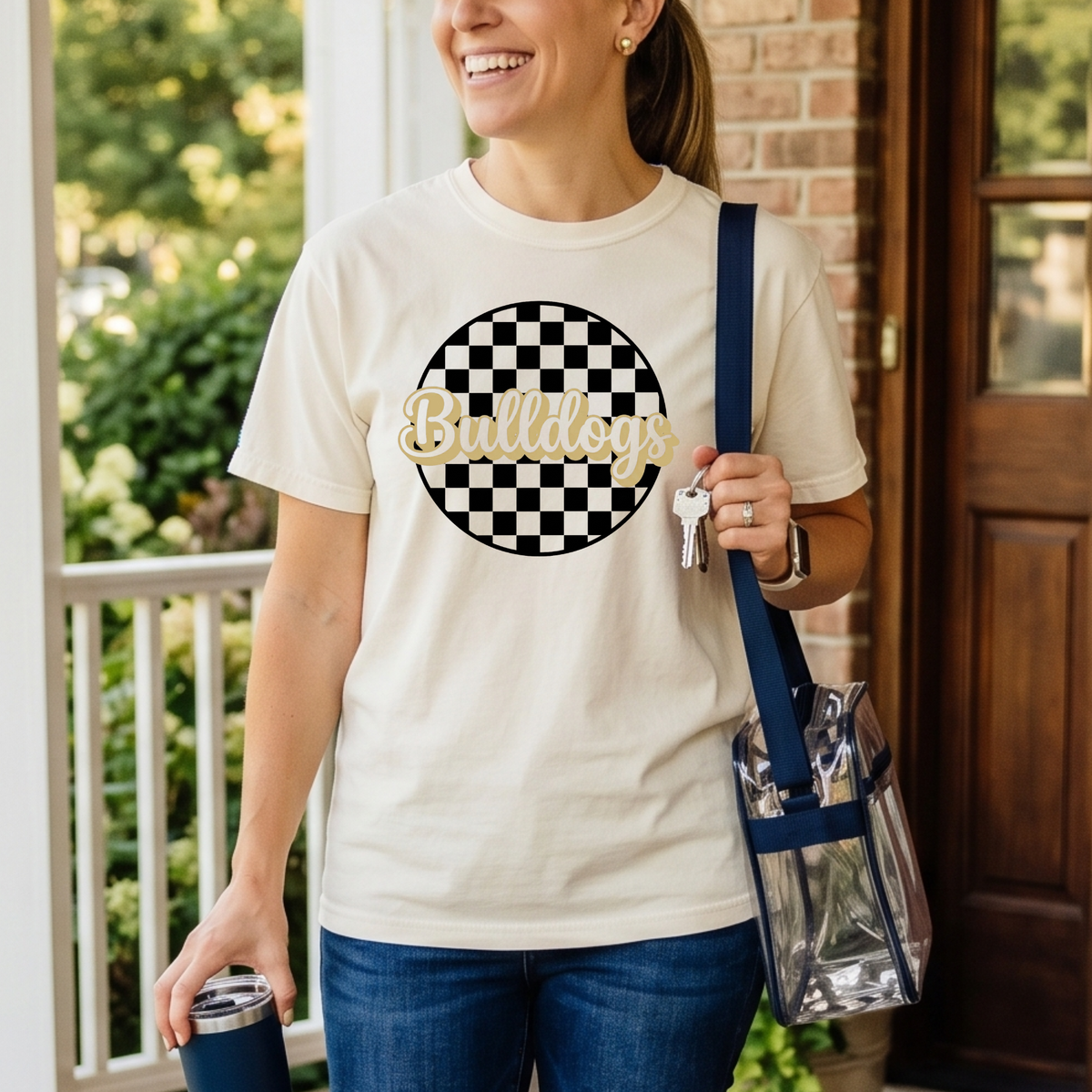 Woman wearing a white t-shirt with a checkered circle design and 'Bulldogs' text, holding a blue bag and a blue tumbler.