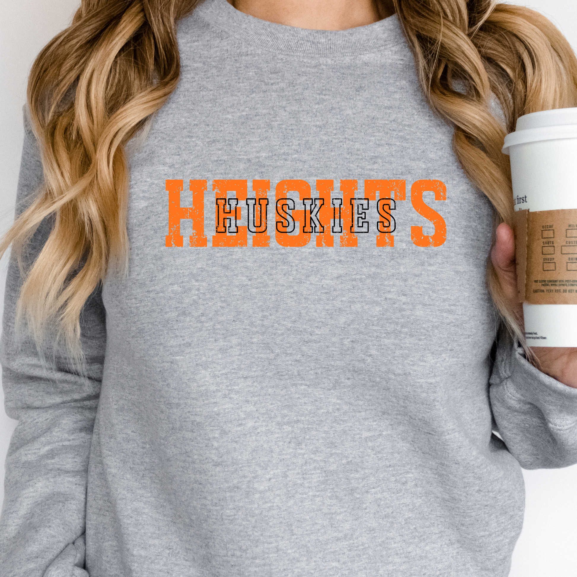 A person wearing a grey crewneck sweatshirt featuring a distressed orange 'HEIGHTS' design and white 'HUSKIES' text layered on top, holding a coffee cup.