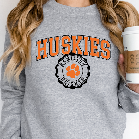 A person wearing a grey crewneck sweatshirt featuring a bold orange 'HUSKIES' arch design and a 'HAMILTON HEIGHTS' circular mascot seal, holding a coffee cup.