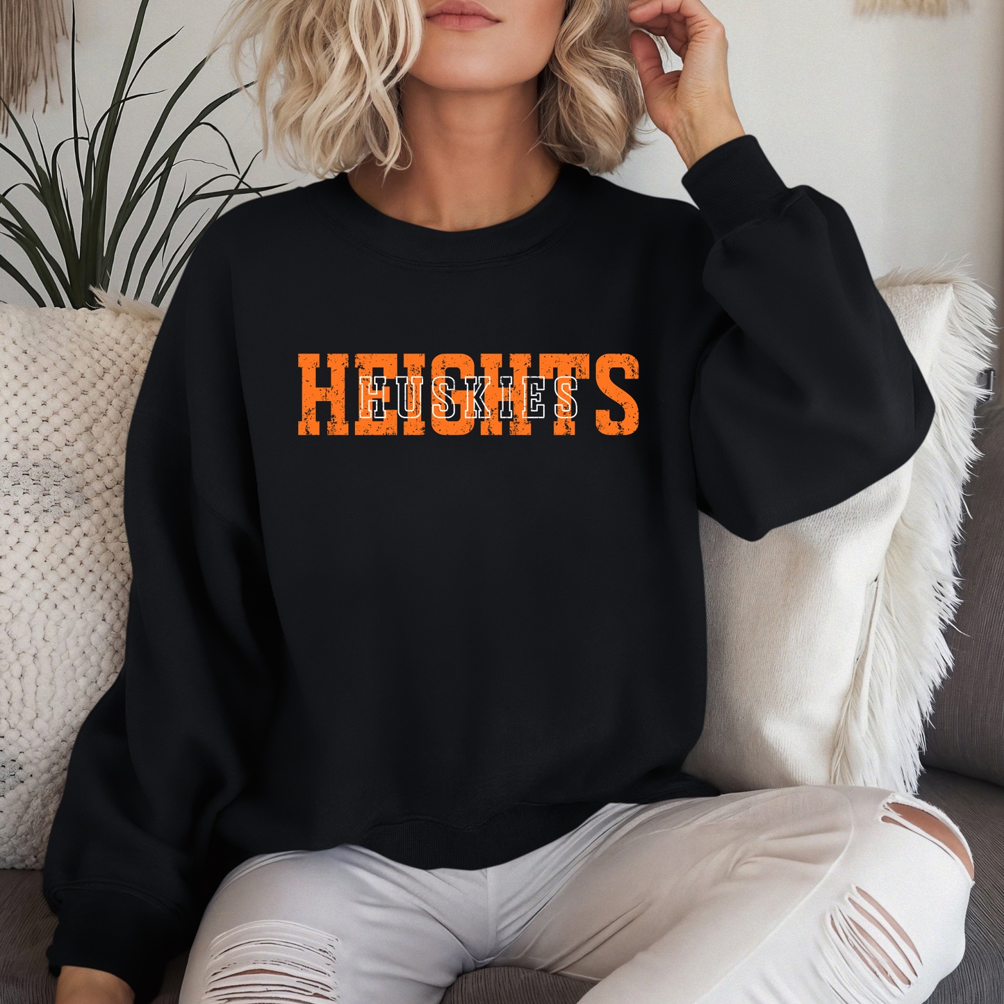 A person sitting on a couch wearing a black crewneck sweatshirt featuring a distressed orange 'HEIGHTS' design with 'HUSKIES' in white block lettering layered on top.