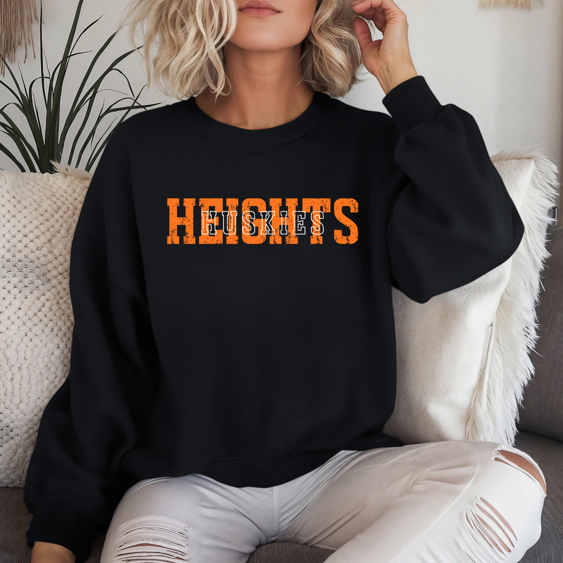 A person sitting on a couch wearing a black crewneck sweatshirt featuring a distressed orange 'HEIGHTS' design with 'HUSKIES' in white block lettering layered on top.