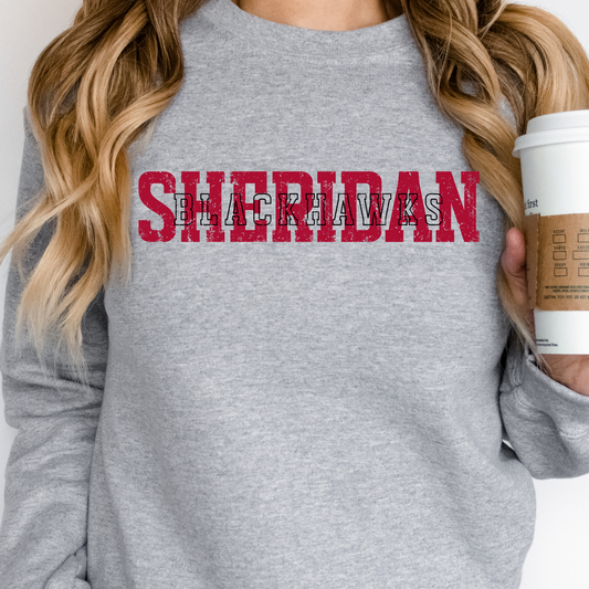 Woman holding a coffee cup, wearing a grey crewneck sweatshirt featuring a layered, vintage-style Sheridan Blackhawks typography graphic.