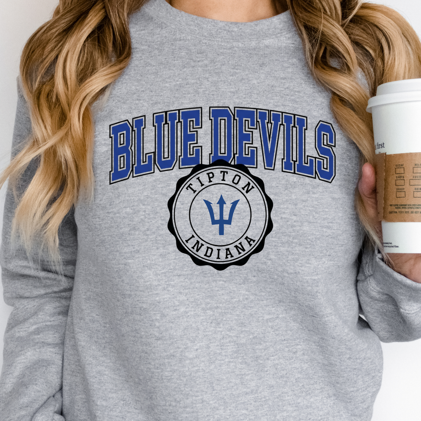 Woman holding a coffee cup, wearing a grey crewneck sweatshirt featuring a collegiate-style Tipton Blue Devils and Indiana seal graphic.