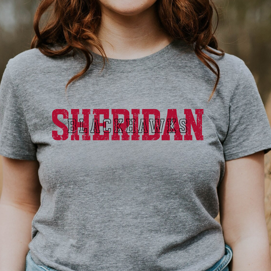 Woman wearing a grey graphic t-shirt featuring a layered, vintage-style Sheridan Blackhawks typography design.
