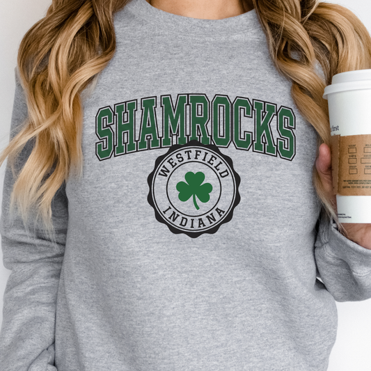 A person wearing a soft grey crewneck sweatshirt featuring a collegiate-style 'SHAMROCKS' arch design positioned above a circular 'WESTFIELD INDIANA' shamrock seal, holding a disposable coffee cup.