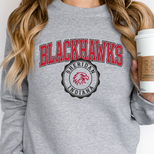 Woman holding a coffee cup, wearing a grey crewneck sweatshirt featuring a classic collegiate Sheridan Blackhawks graphic and team seal.