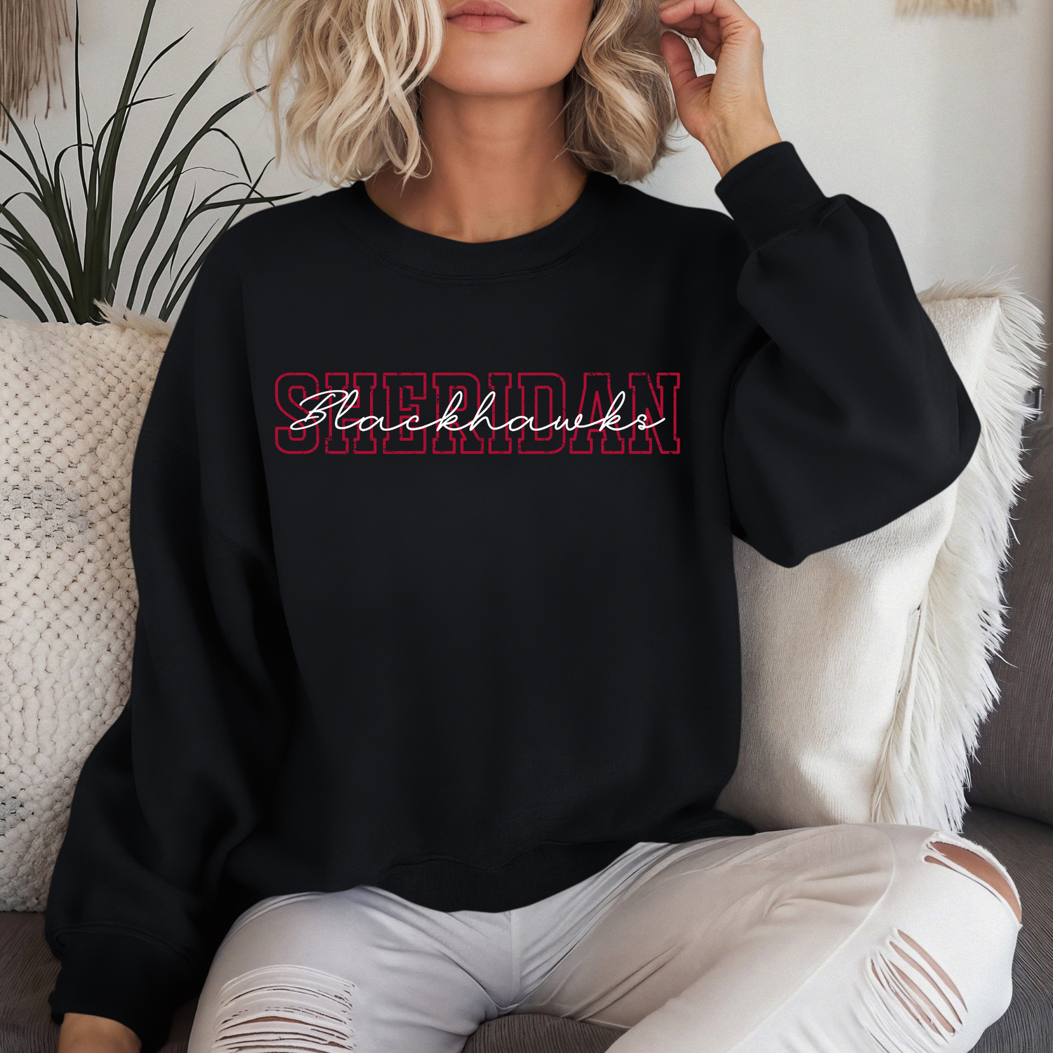 Woman wearing a black crewneck sweatshirt featuring a layered, vintage-style Sheridan Blackhawks block and script typography design, sitting on a couch.