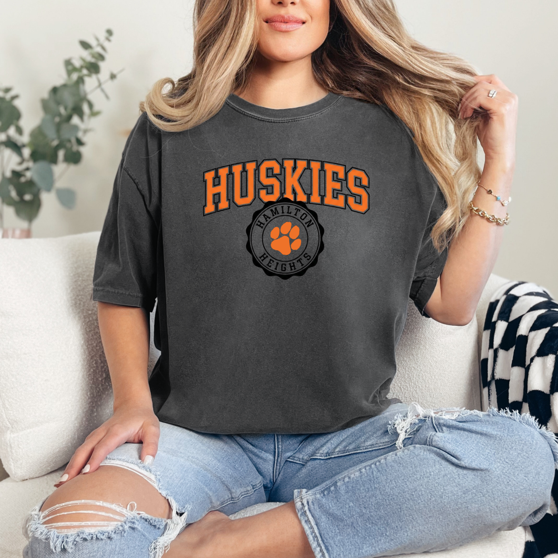 A person wearing a charcoal-colored graphic tee featuring a bold orange 'HUSKIES' arch design and a 'HAMILTON HEIGHTS' circular mascot seal.