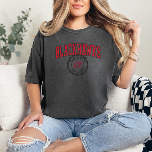 Woman wearing a dark charcoal t-shirt with a vintage-style Sheridan Blackhawks collegiate design, sitting on a couch.