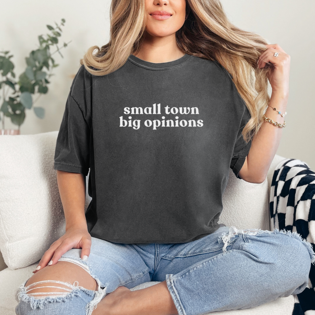 Woman sitting on a sofa wearing a Pepper graphic tee with the phrase ‘small town big opinions’; casual Midwest-inspired outfit with ripped jeans.