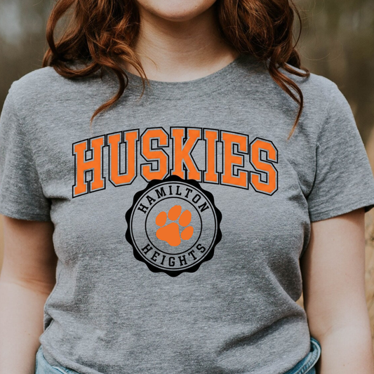 A person wearing a heather grey graphic tee featuring a bold orange 'HUSKIES' arch design and a 'HAMILTON HEIGHTS' circular mascot seal.